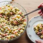 Olivier Salad from Russia: 5 Proven Secrets for a Perfect Dish Top-down view of a crystal bowl of creamy Olivier salad from Russia with a silver serving spoon held by a hand.